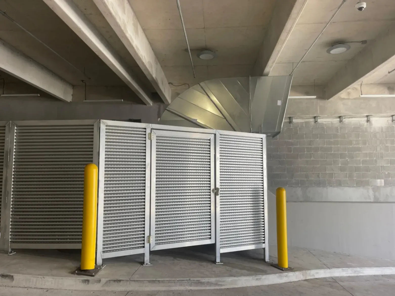 Custom Metal Fabrication in Deerfield Beach: Silver metal louvered panels acting as a security enclosure in a concrete parking structure. The setup features an integrated hinged gate with a latch, a large overhead galvanized duct, and two protective yellow steel bollards in the foreground.