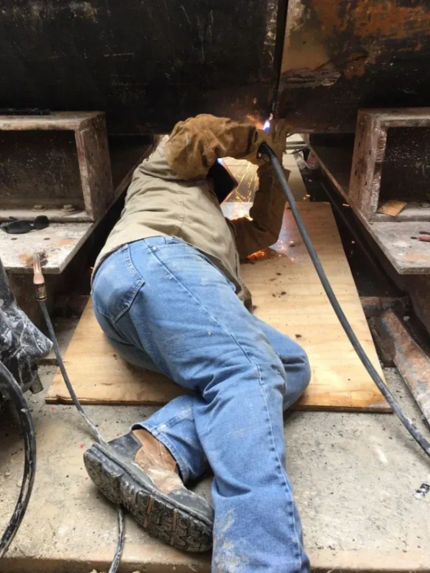 A technician from Abel Welding Services lies on a wooden board to perform precise heavy fabrication welding underneath a large industrial structure in Davie, FL.