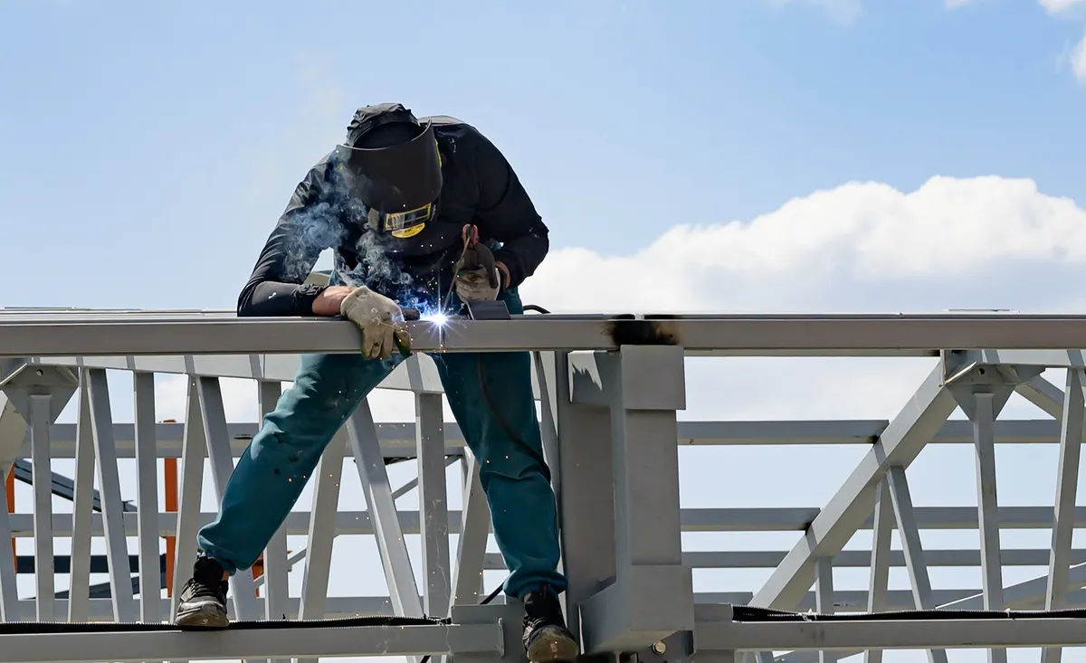 Abel Welding Services offers elite structural steel welding in Deerfield Beach, FL. An individual wearing a black long-sleeved shirt, dark pants, heavy-duty gloves, and a protective welding helmet with a yellow visor, working on a large, grey metal structure. Smoke and bright sparks are visible near their hands as they weld a seam.
