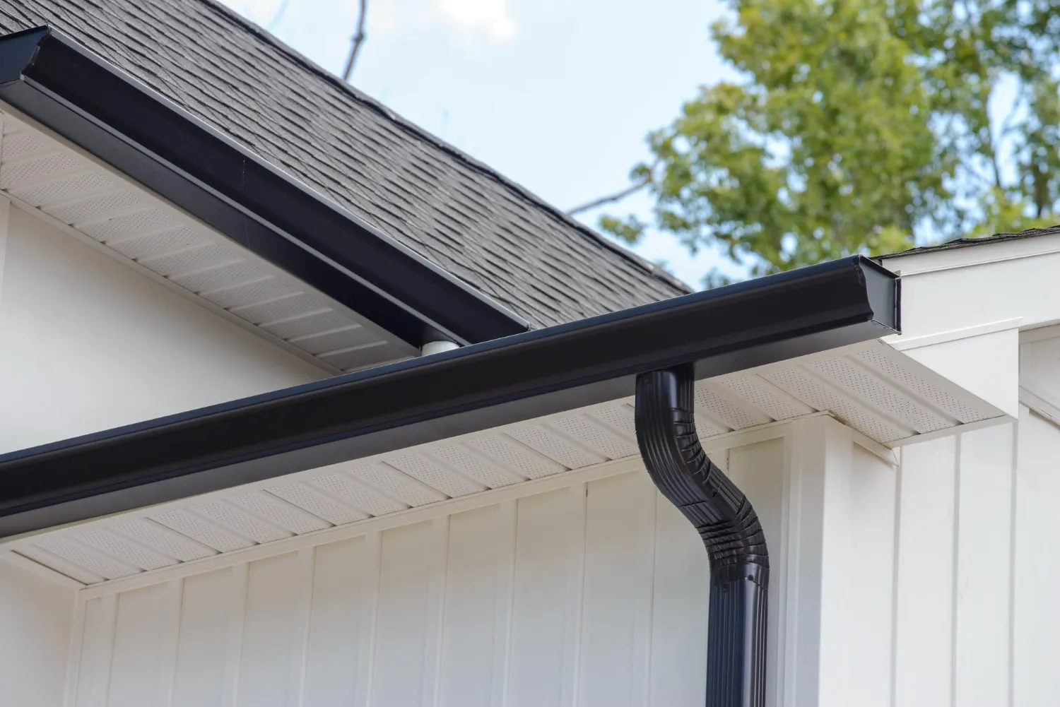 A close-up view of a sleek, black seamless gutter installation in Manchester, NH, featuring a matching downspout and clean white siding accents. This professional drainage system by JM Exteriors Roofing Siding prevents costly water damage by diverting rain away from the home's foundation and siding.