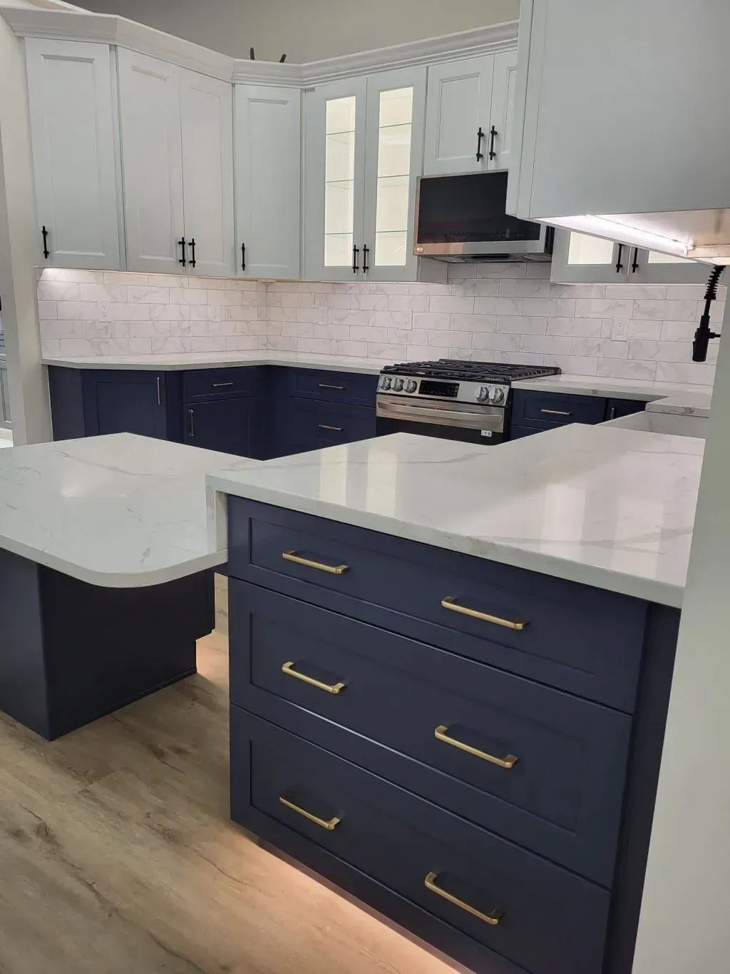 Two-tone custom cabinet installation for a kitchen remodel in Fairfax, VA, showcasing deep navy blue base drawers with gold hardware, white upper shaker cabinets with glass door inserts, and white quartz countertops with under-cabinet lighting.