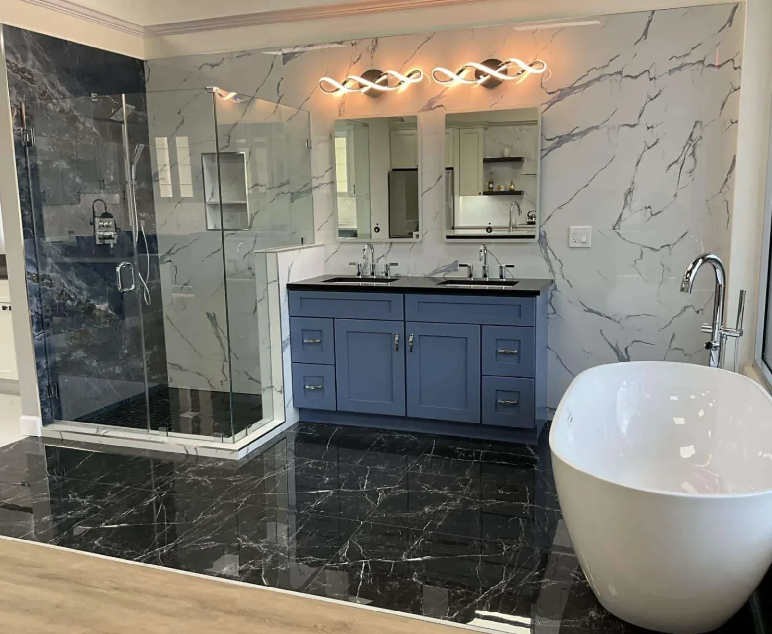 A luxury bathroom remodeling in Fairfax, featuring highly polished black marble floor tiles, a dusty blue double vanity with a black countertop, and a white freestanding soaking tub. The back wall is covered in white marble-look tile, showcasing two smart mirrors under wavy LED light fixtures next to a frameless glass walk-in shower.