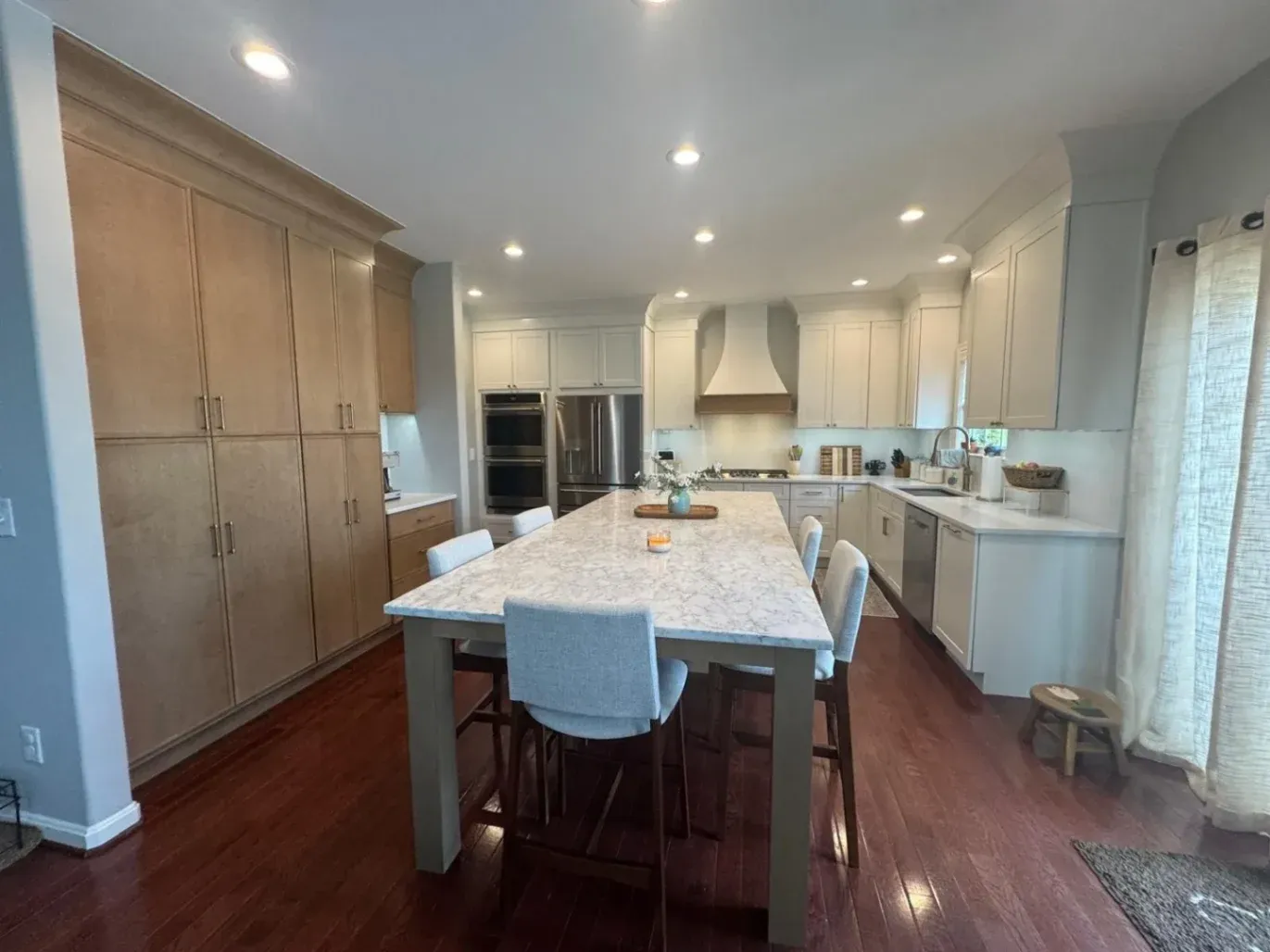 A luxury open-concept kitchen remodel near Fairfax, VA, completed by KBA Kitchen and Bath Design and Build, featuring a grand marble-look quartz island with seating, premium hardwood floors, custom off-white cabinetry, and a massive natural wood pantry wall.