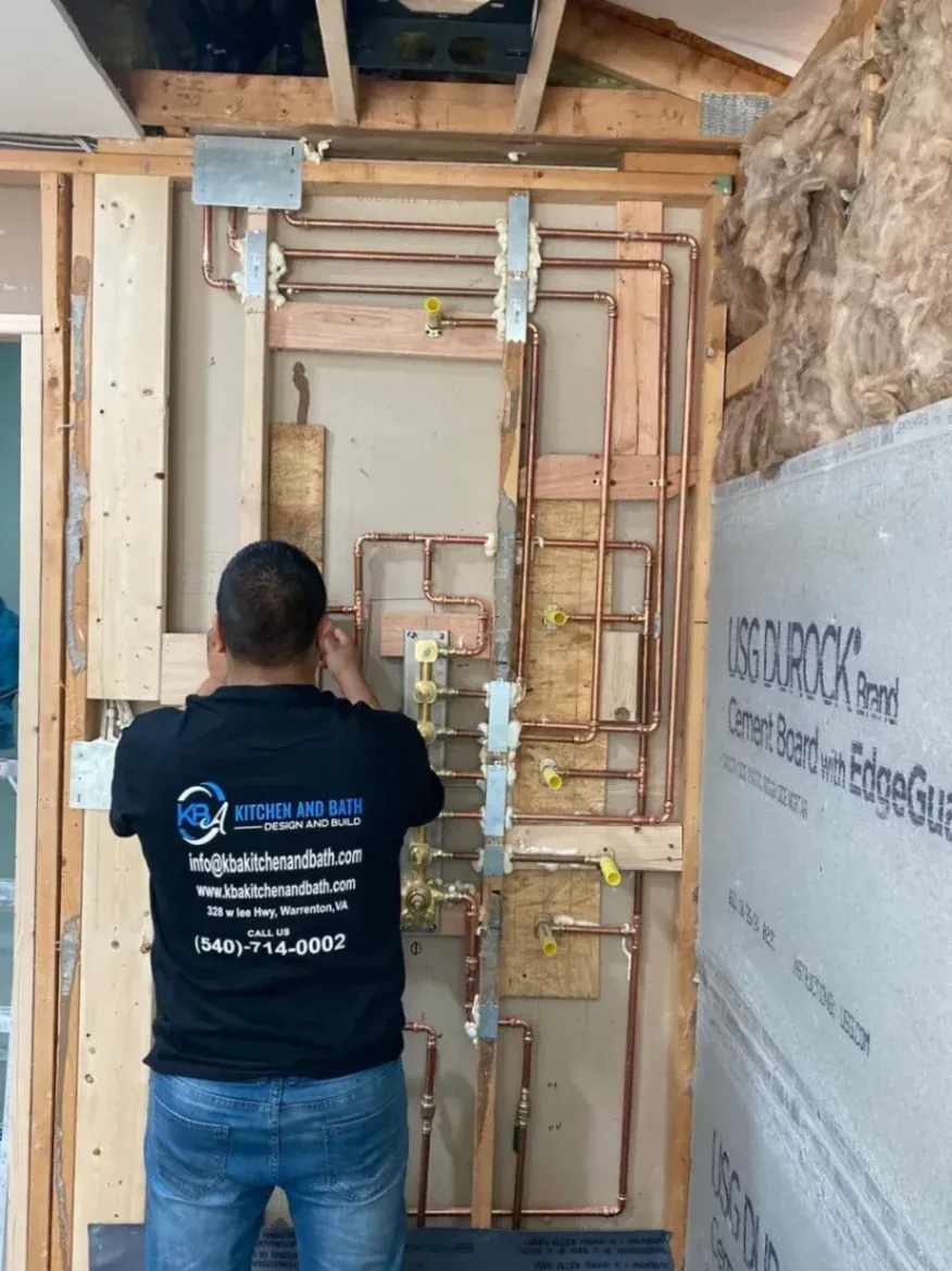 A professional contractor seen from behind wearing a black KBA Kitchen and Bath t-shirt, working on a complex network of newly installed copper plumbing lines inside an exposed wooden wall frame during a bathroom renovation in Fairfax, VA.