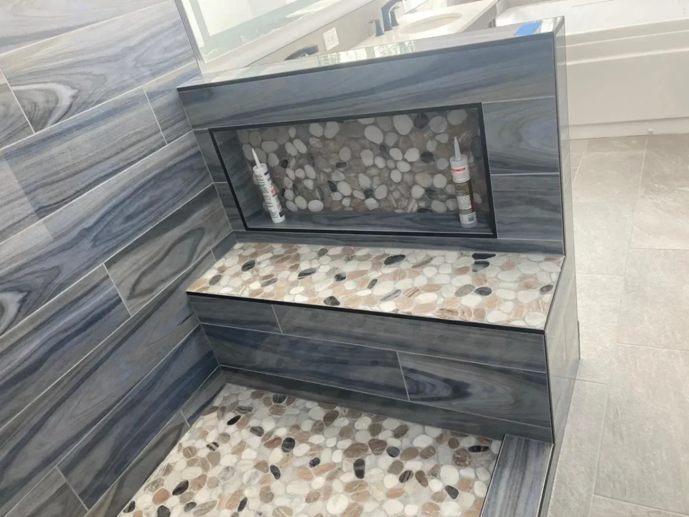 Durable custom shower tile installation in Fairfax by KBA Kitchen and Bath, showcasing a natural pebble stone shower floor, a matching built-in bench, and high-quality blue-gray wood-grain wall tiles built to last.