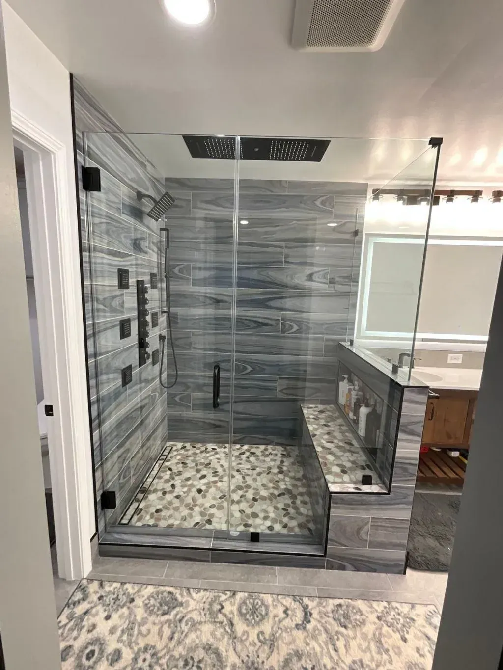 A modern luxury walk-in shower with a frameless glass enclosure, featuring gray and blue wood-look wall tiles, a natural pebble stone floor, a matching built-in shower bench, and premium matte black shower fixtures with body jets. This is the result of a bathroom remodeling in Fairfax, VA.