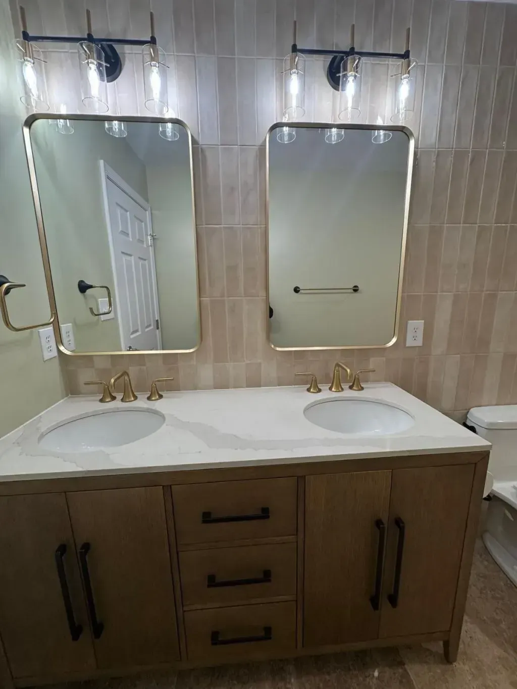 A bathroom remodeling in Fairfax featuring a double vanity with a natural wood cabinet with modern black handles and a white quartz countertop with soft gray veining. Two undermount sinks are paired with brushed gold widespread faucets. On the wall, two rounded rectangular mirrors with gold frames are mounted against a backdrop of vertically stacked neutral beige subway tiles, lit by contemporary black-and-brass light fixtures.