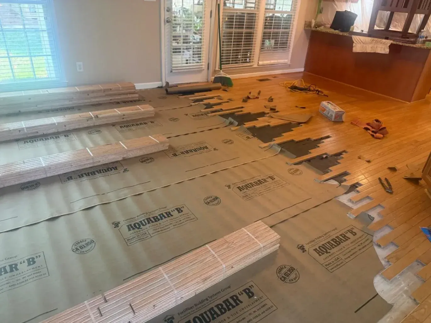 In-progress hardwood floor installation in a Clayton, NC home by DNH Hardwood Flooring LLC. The photo shows protective AQUABAR 'B' underlayment rolls and stacks of new wood planks ready for layout.
