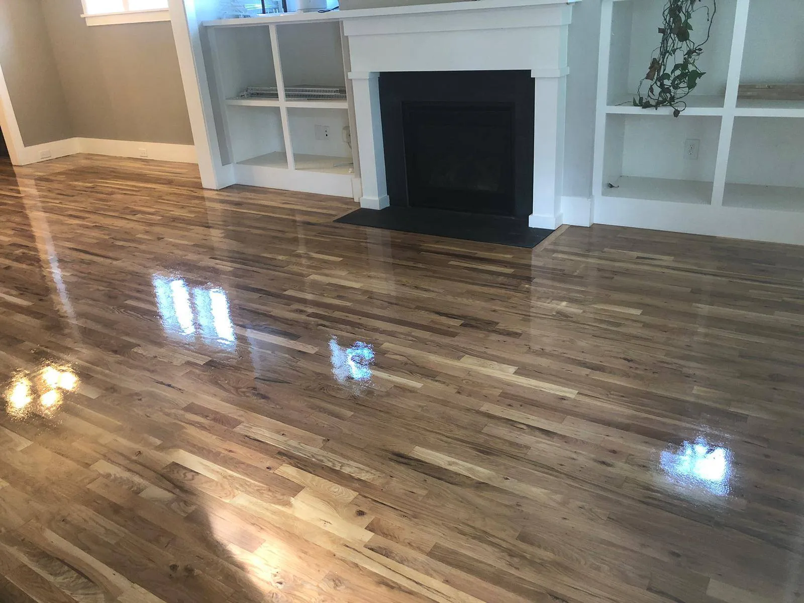 Freshly refinished high-gloss hardwood floors in Raleigh. The floors are reflecting window light in a living room with a white fireplace and built-in shelving.