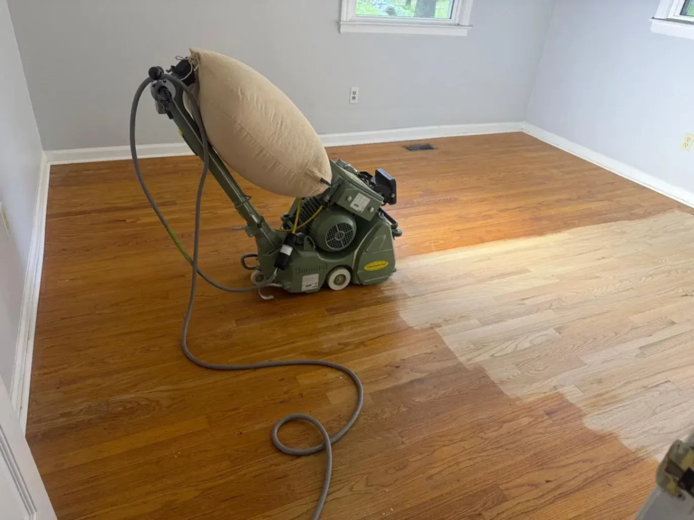 DNH Hardwood Flooring using a heavy-duty belt floor sander for sanding and refinishing services in Raleigh, NC. The sander is resting on a hardwood floor mid-sanding process, showing raw bare wood contrasting against the darker old finish.