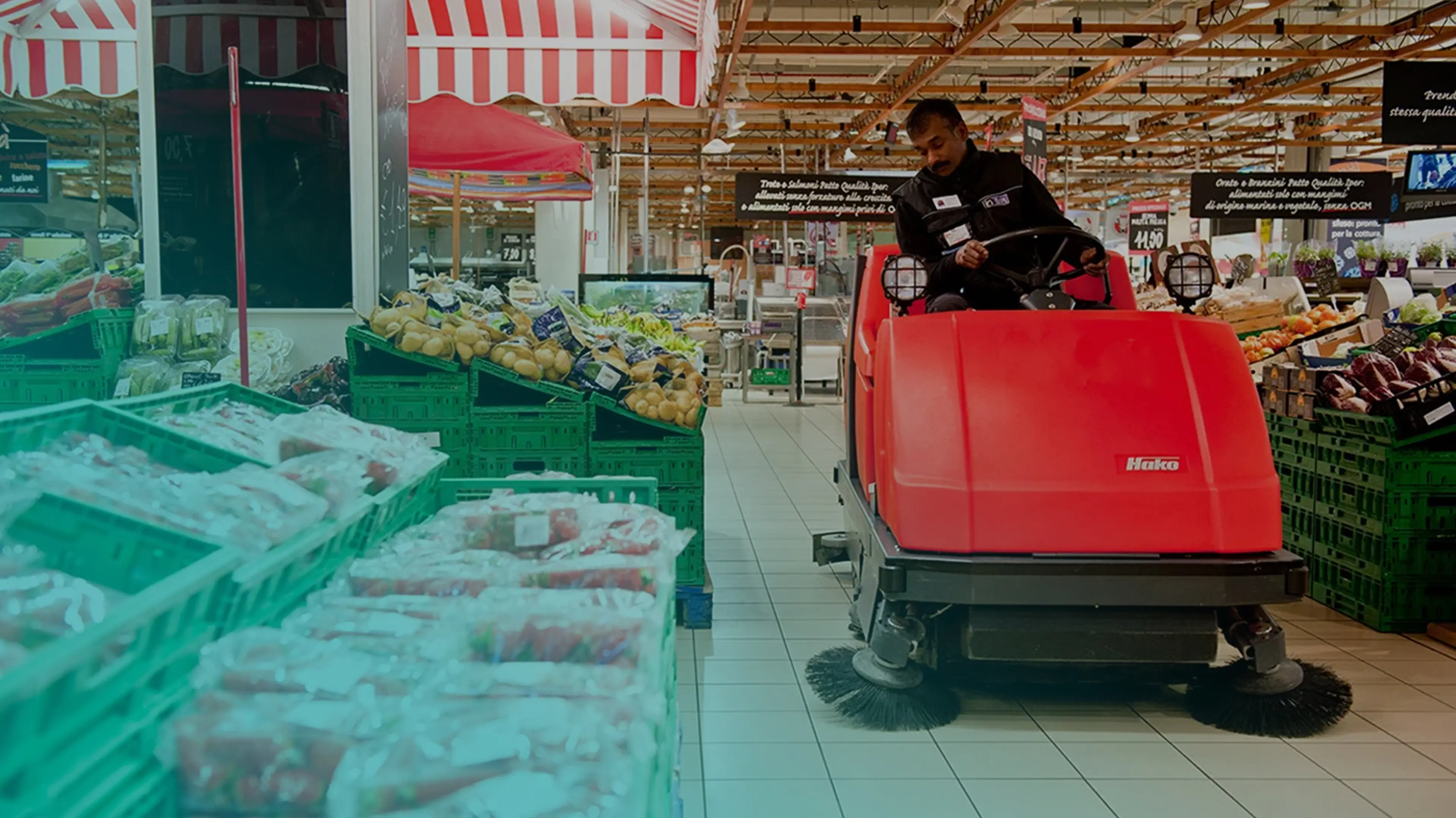 Un addetto alle pulizie usa una spazzatrice Hako in un supermercato vicino a verdure e frutta.