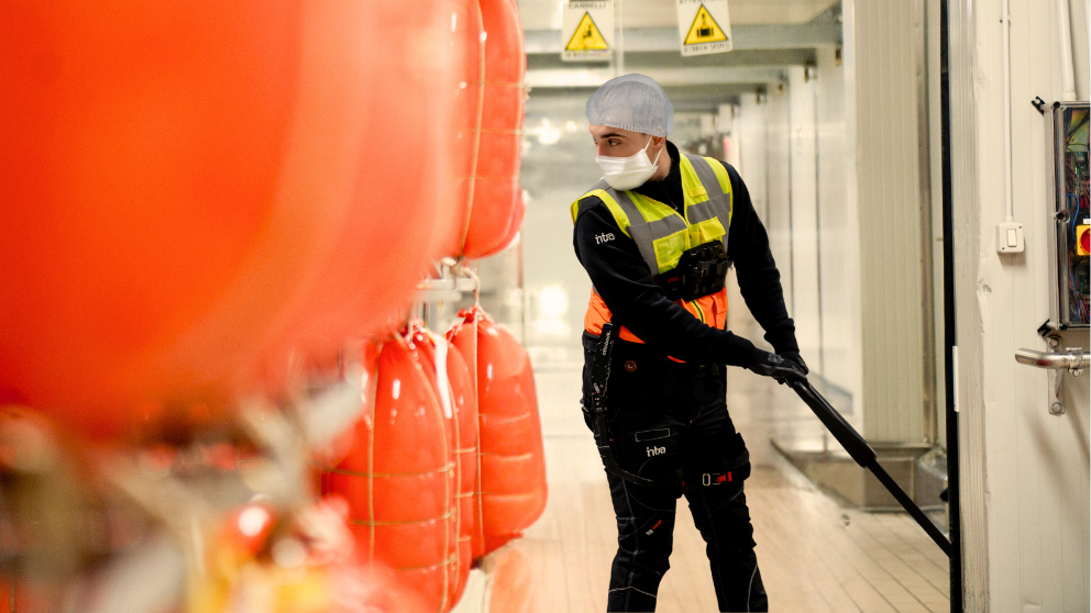 Ecco una possibile didascalia per l'immagine:

Un addetto alla sanificazione che indossa un DPI sta pulendo in un impianto di trasformazione alimentare. L'immagine è scattata con il personale che lavora dietro i sacchi di cibo in un corridoio.