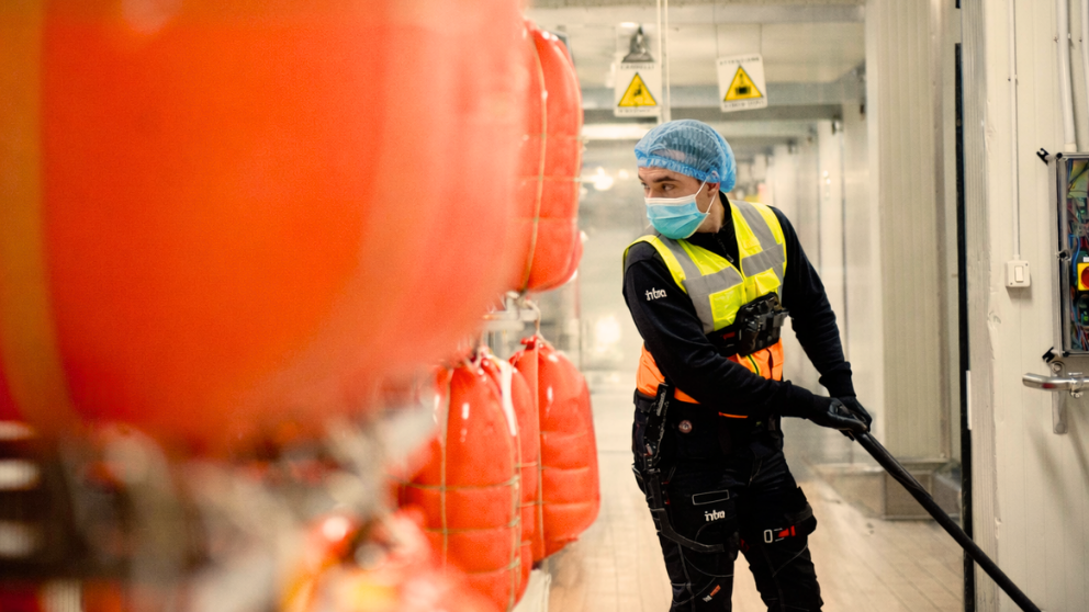 Ecco una possibile didascalia per l'immagine:

Un addetto alla sanificazione che indossa un DPI sta pulendo in un impianto di trasformazione alimentare. L'immagine è scattata con il personale che lavora dietro i sacchi di cibo in un corridoio.