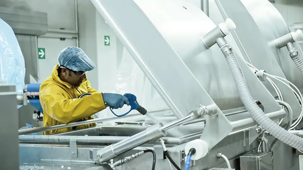 Ecco una didascalia per l'immagine:

"Un uomo che indossa un impermeabile giallo, un cappello e dei guanti sta pulendo una macchina industriale argentata in un ambiente commerciale igienico."