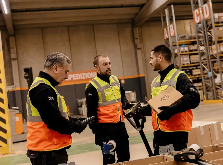 Tre lavoratori in magazzino indossano giubbotti di sicurezza mentre discutono.