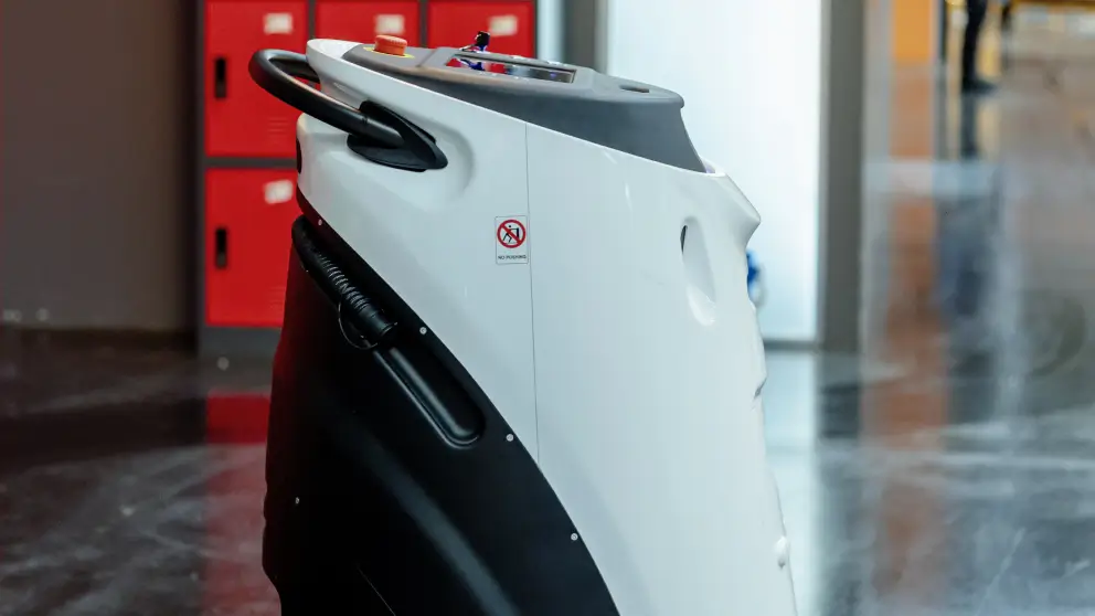 Ecco una didascalia per l'immagine:

Un robot aspirapolvere autonomo bianco si trova in un corridoio con gli armadietti rossi sullo sfondo. Il robot ha un display nella parte superiore e un pulsante rosso di arresto di emergenza. Sul lato del robot è presente un adesivo con la scritta "no pushing".