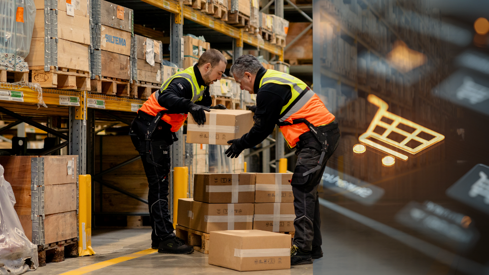 Due dipendenti di un magazzino indossano giubbotti di sicurezza e lavorano insieme per impilare le scatole. Le scatole sono impilate su un pallet. Sul lato destro dell'immagine è presente un'immagine di un carrello della spesa per rappresentare il commercio elettronico. Lo sfondo è un magazzino pieno di ripiani e oggetti confezionati.
