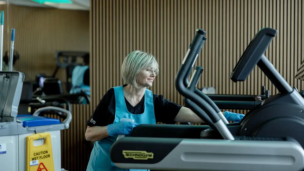 Ecco una possibile didascalia per l'immagine:

In questa immagine, una donna indossa un grembiule blu e guanti usa e getta blu mentre pulisce una macchina per esercizi Technogym con una bottiglia spray e uno straccio. La donna sorride mentre pulisce la macchina per esercizi in una palestra. Sullo sfondo, si vede un cartello di avvertimento di pavimento bagnato.