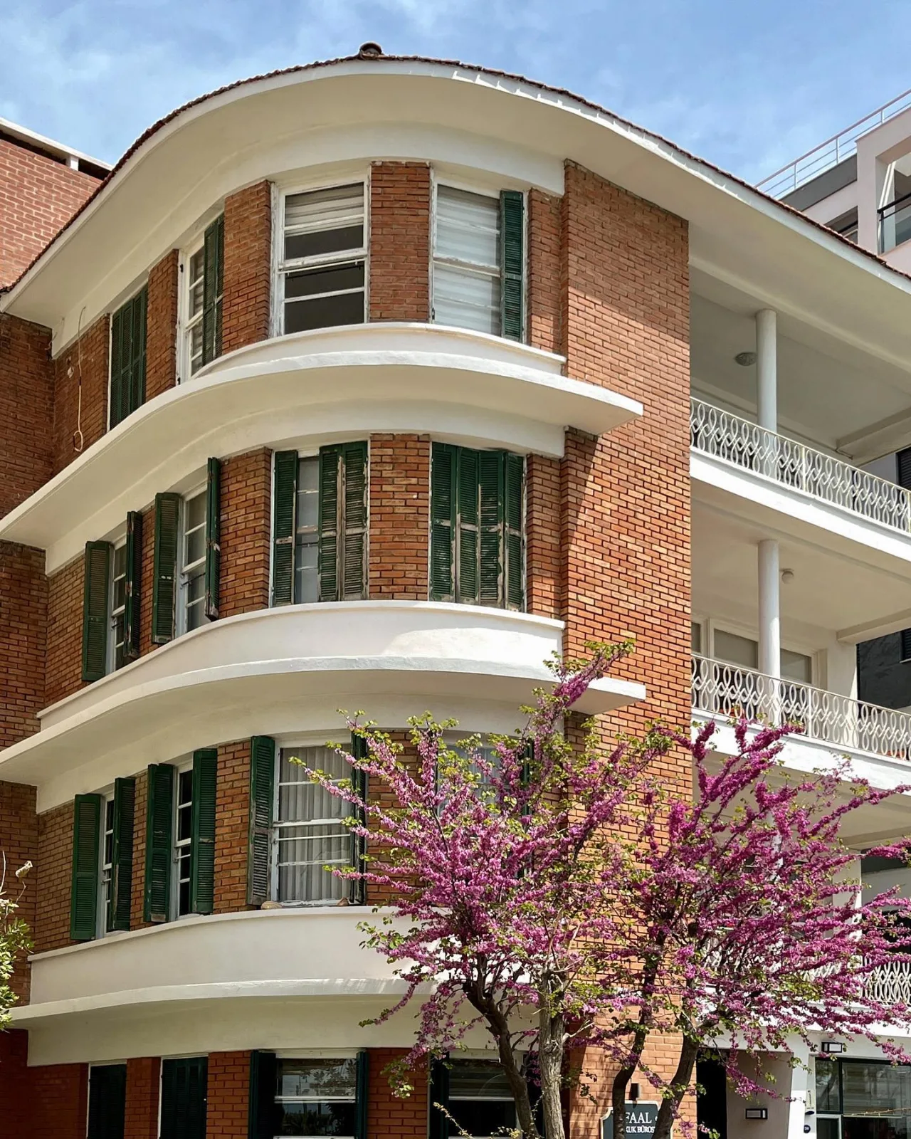 Paya Apartmanı’nın eğrisel köşe cephesi; kırmızı tuğla yüzeyler, yeşil ahşap kepenkler, beyaz yatay bordürler ve çiçekli bir ağaç görülüyor.
