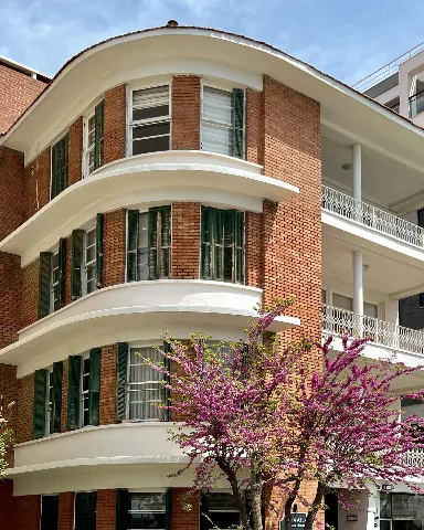 Paya Apartmanı’nın eğrisel köşe cephesi; kırmızı tuğla yüzeyler, yeşil ahşap kepenkler, beyaz yatay bordürler ve çiçekli bir ağaç görülüyor.