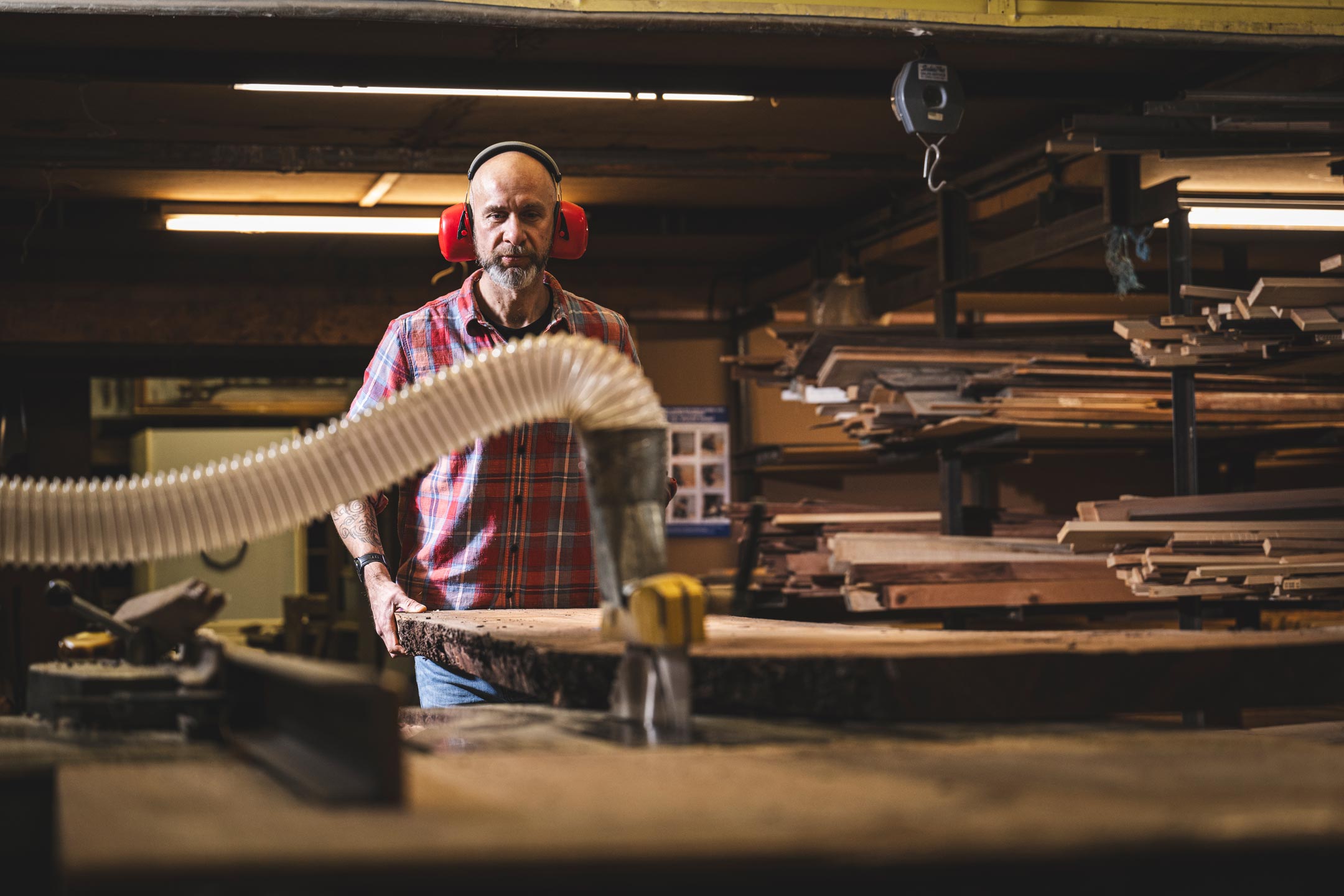 Person wearing ear protection in a woodshop with tools and stacked wooden planks.