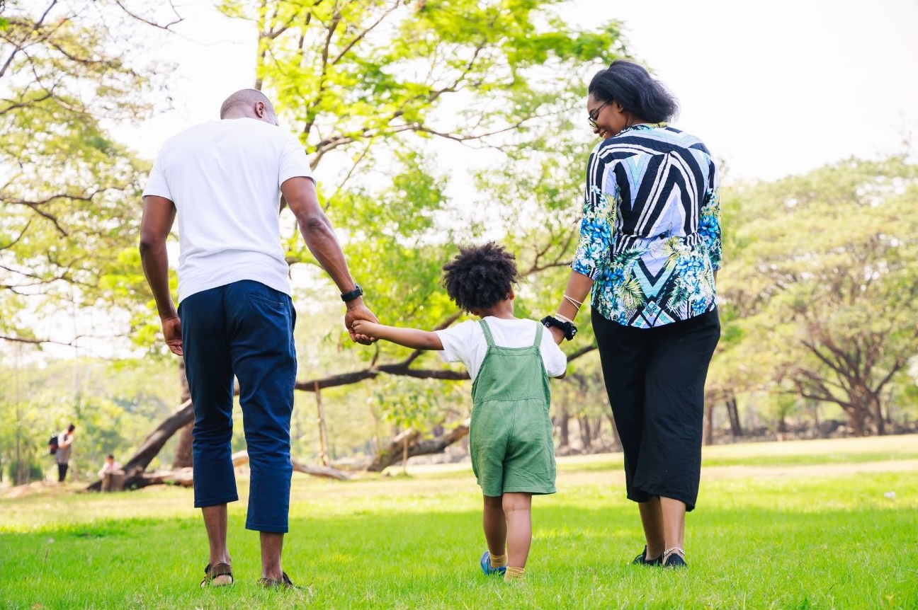 Two adults and a child holding hands, walking away across a sunny park lawn