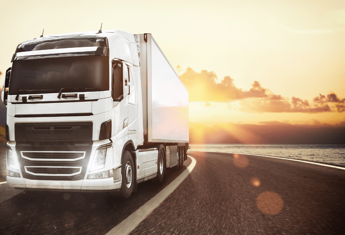 A white freight truck driving on a curved coastal road at sunset, symbolising the efficiency and reliability of freight haulage companies in delivering goods over long distances.