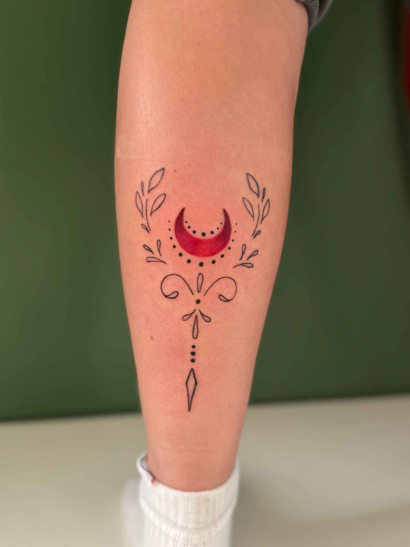 Tatouage fin et symétrique représentant un croissant de lune rouge au centre, entouré de motifs végétaux délicats et de 