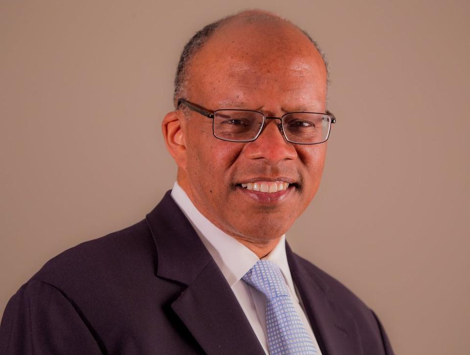 Howard Wooley posing in a white shirt and light blue tie and black suitjacket. He wears rectangular glasses.
