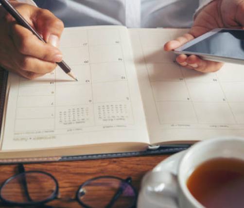 A person updating their daily journal planner. The person is holding their phone in one hand and a pencil in the other. They are sitting on a table, which shows a pair of glasses and a cup of tea
