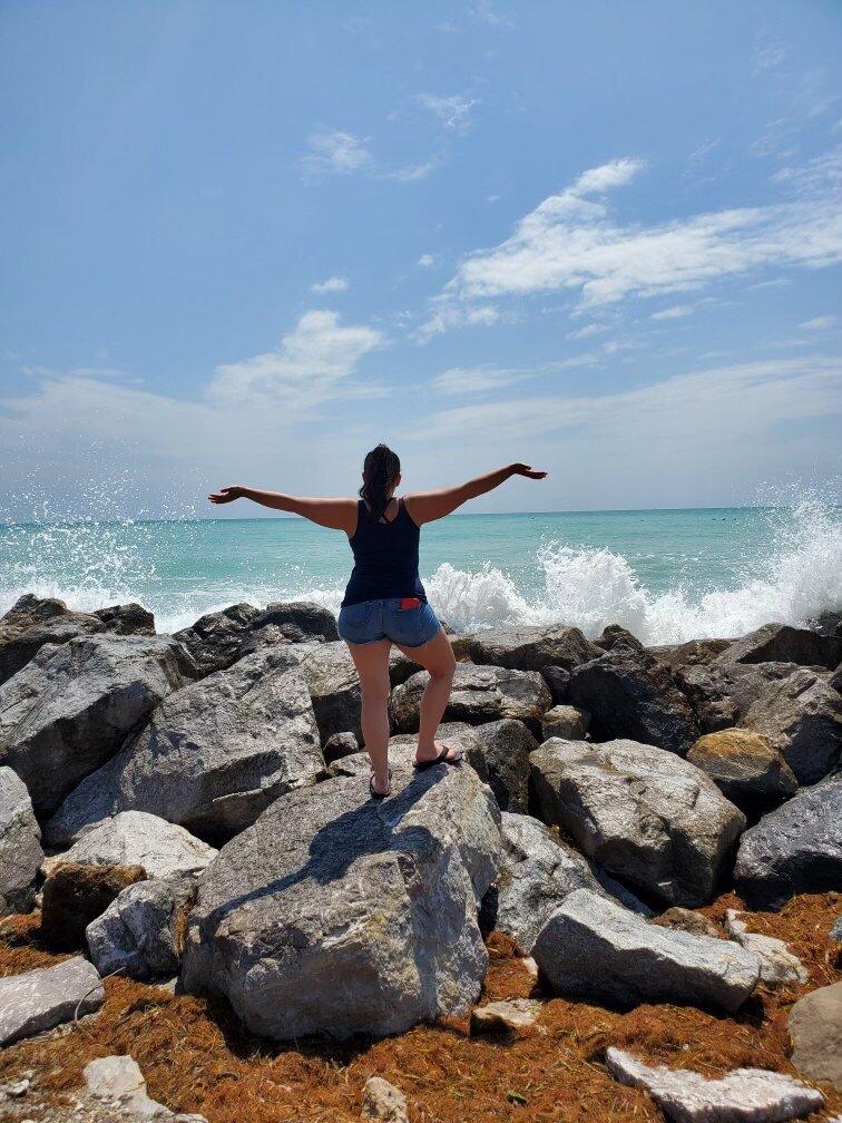 Linsday, facing the sea, arms outstretched in triumphant victory after finding her path toward stability and healing while living with MG