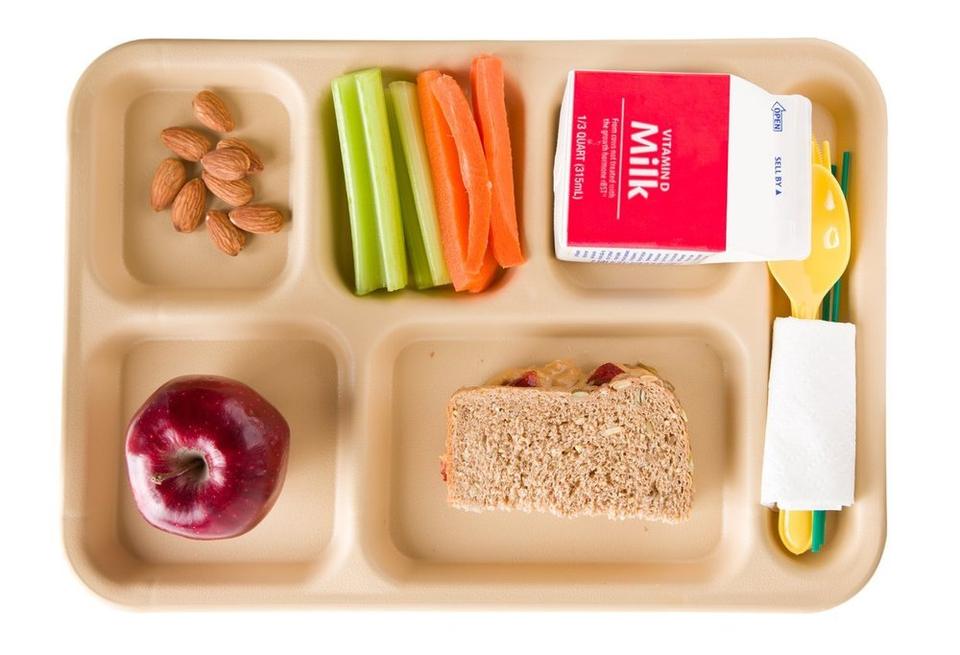 A beige lunch tray featuring almonds, cut carrots and celery, a Vitamin D fortified milk, a red apple, and a half of a peanut butter and jelly sandwich, with some cutlery and a napkin