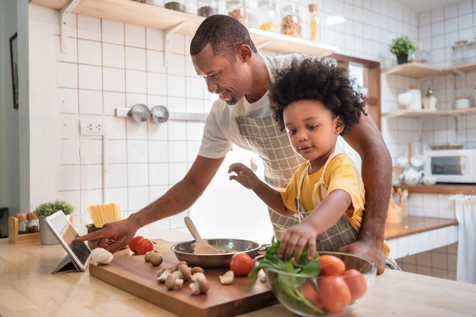 A father and child are cooking together in the kitchen. The father is using an ipad and the child is selecting produce from a glass bowl.