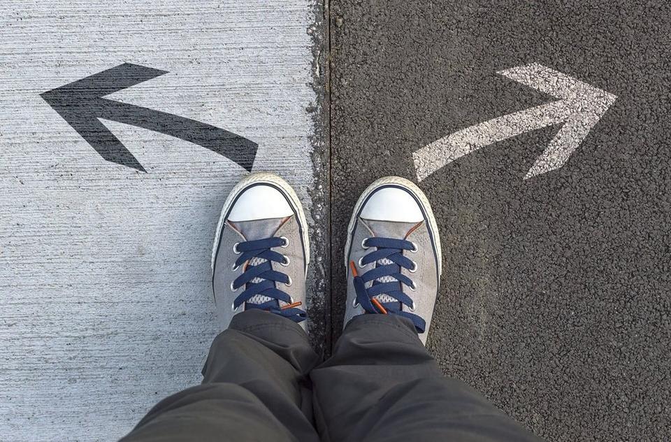 A closeup of a person's feet wearing sneakers and deciding what path to take. The 2 potential paths are suggested through two different color arrows in opposite directions, by a clear divide in the ground and two different colors of cement