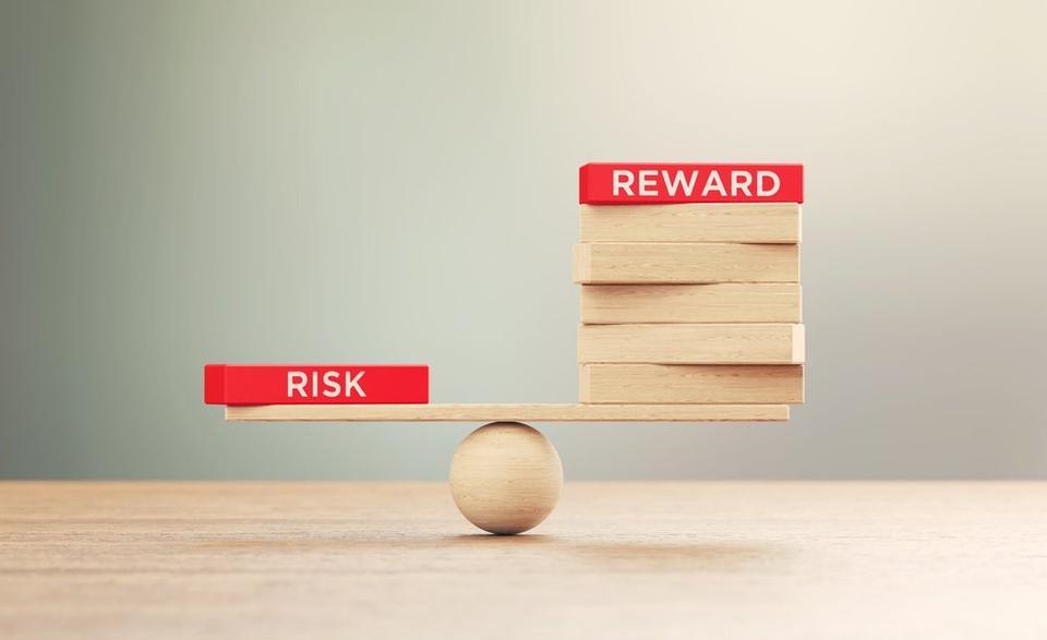 Wooden balance scale with the word 'Risk' on a lower, lighter side and 'Reward' on a higher, heavier side, symbolizing the concept of weighing risks against rewards.