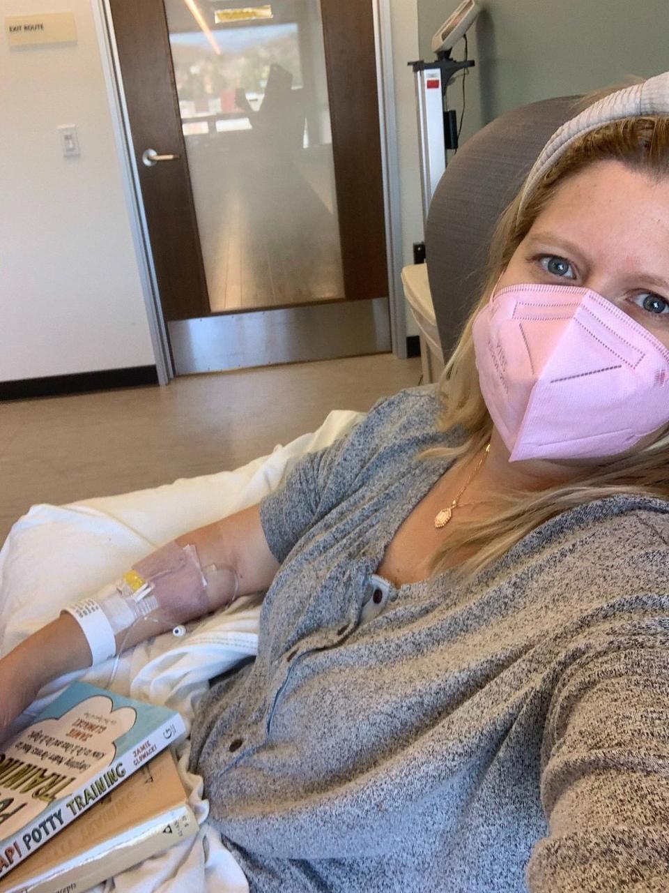 Cate wearing a pink mask and holding books as she receives her infusion