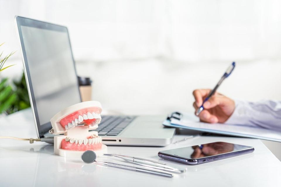 A dental model with teeth placed on a desk next to a laptop, dental tools, and a smartphone, with a person in the background writing on a clipboard.