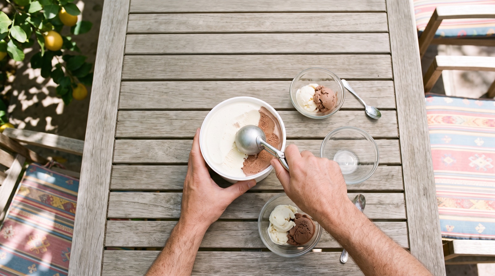 Manos de un adulto sirven helado de vainilla y chocolate desde un envase grande en una mesa de patio con un limonero