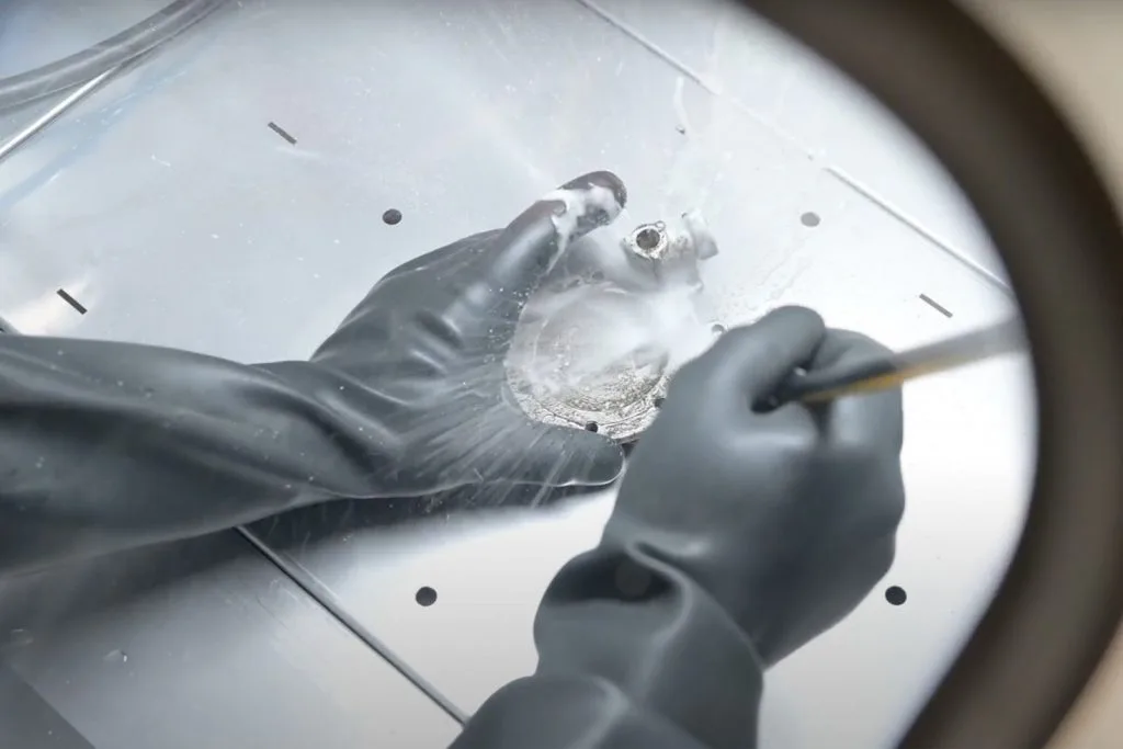 Hands in thick gloves spraying a small metal part through the glove port of a washing booth