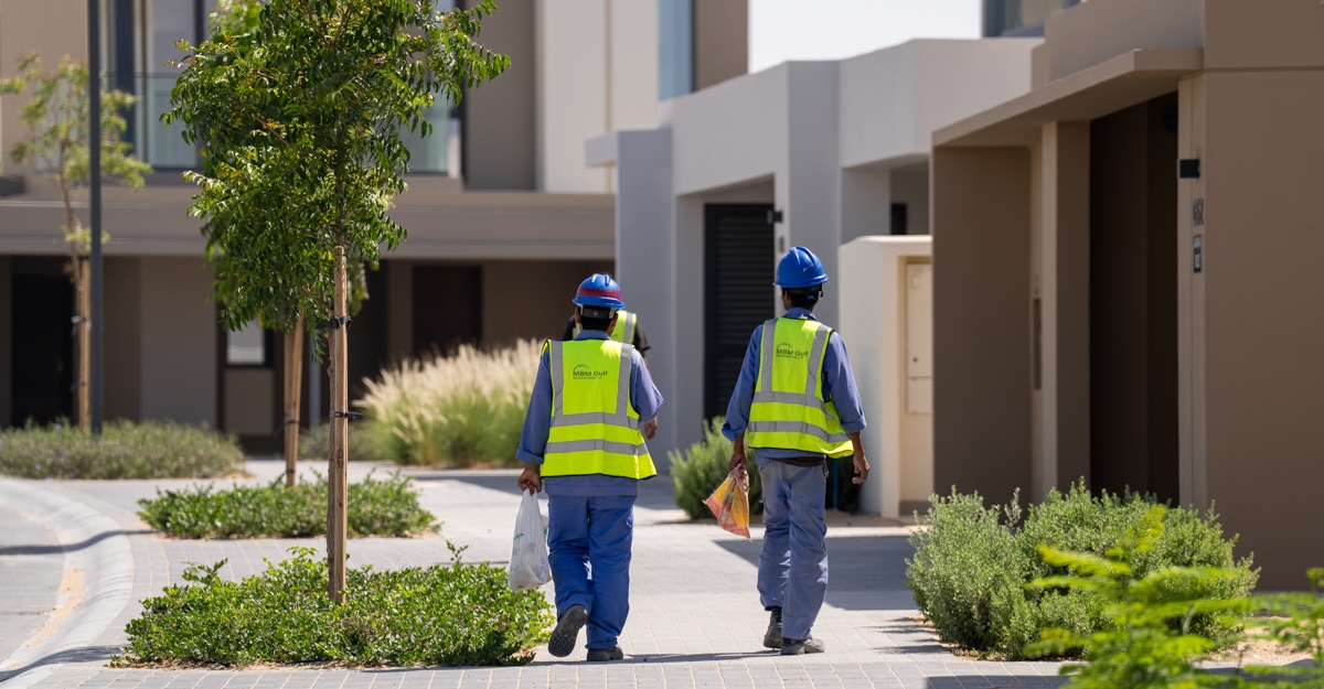 Official Nakheel photograph of Jebel Ali Village villas — two workers in high-visibility vests walking a paved cluster path between newly finished beige homes