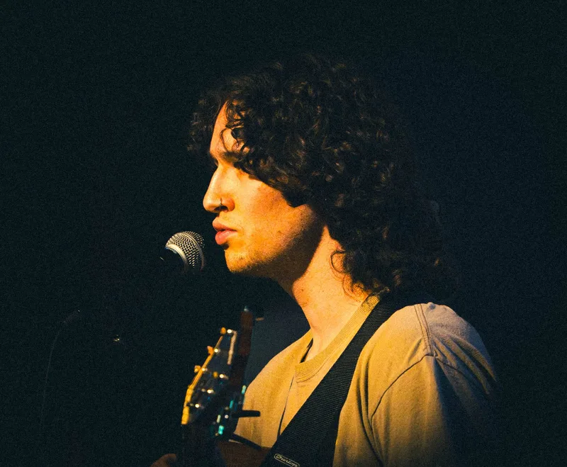 A singer with dark curly hair at a microphone, lit warm gold against a black stage.