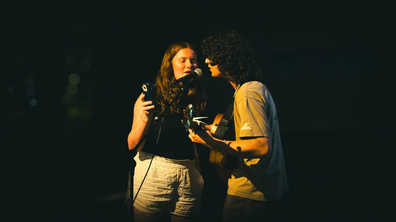 Two performers pressed close at a single microphone, the guitarist mid-strum.