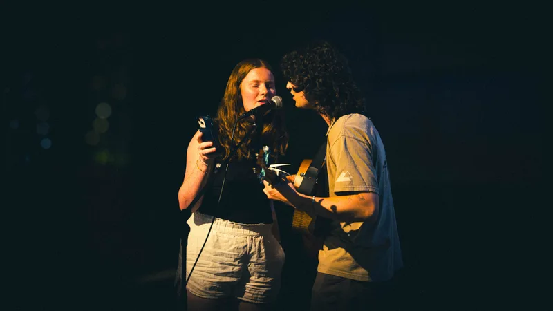 Two performers sharing one microphone, a singer holding a phone and a guitarist leaning in close.