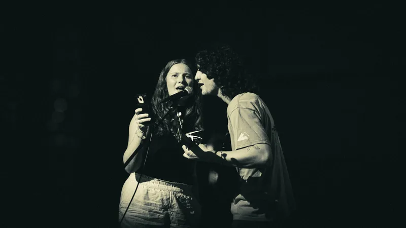 Black and white frame of two singers sharing a microphone, faces almost touching.