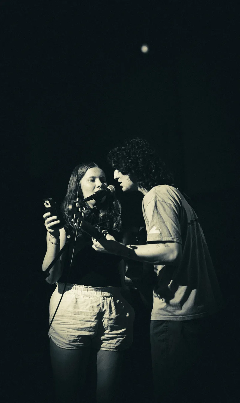 Two performers at a single microphone, shot from a distance in black and white.