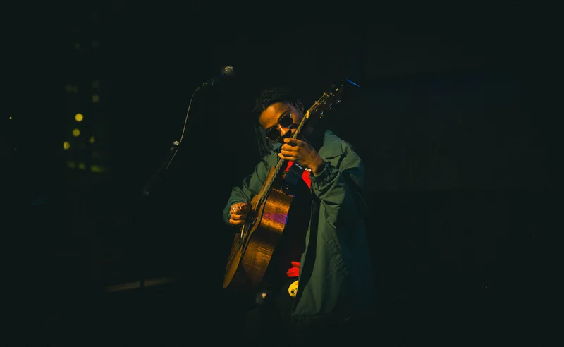 A guitarist in round sunglasses and an oversized jacket, lit from the side on a dark stage.