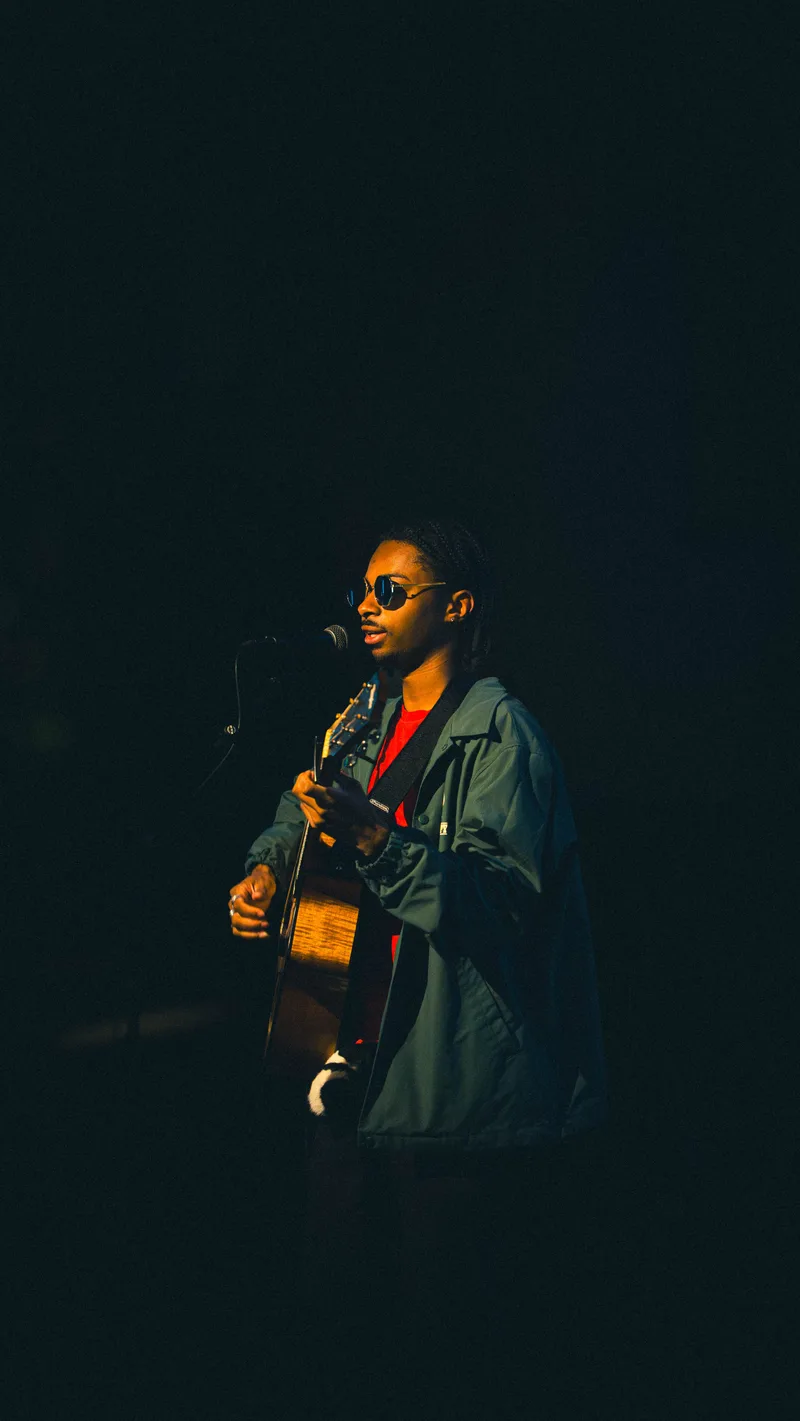 A guitarist in a green jacket sings, one hand shaping a chord on the fretboard.