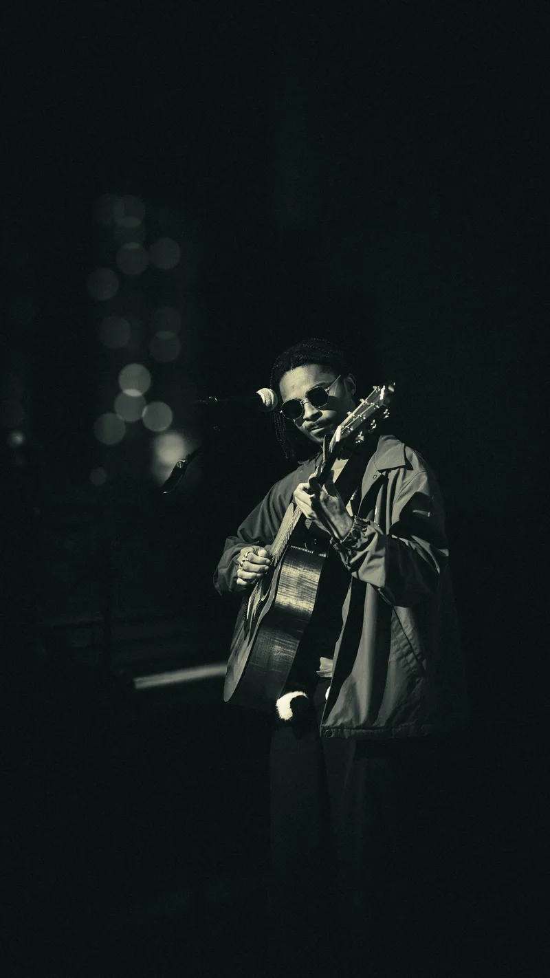 A guitarist looks toward the camera over the neck of his guitar, stage lights blurred behind him.