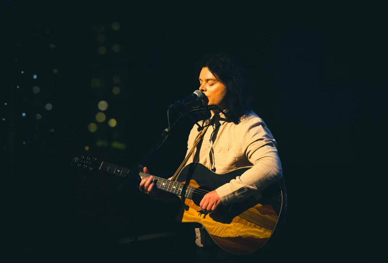 A singer in a linen shirt plays an inlaid acoustic guitar under warm light.
