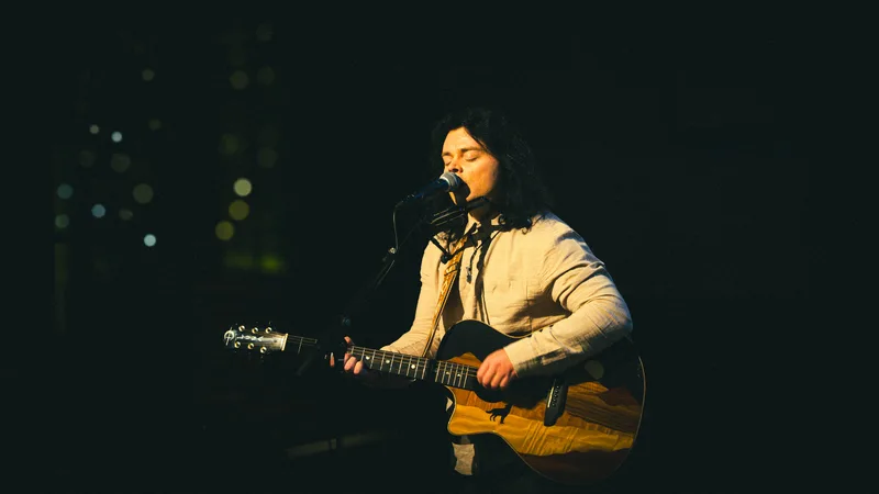 A singer mid-phrase at the microphone, warm light catching the inlay on his guitar.